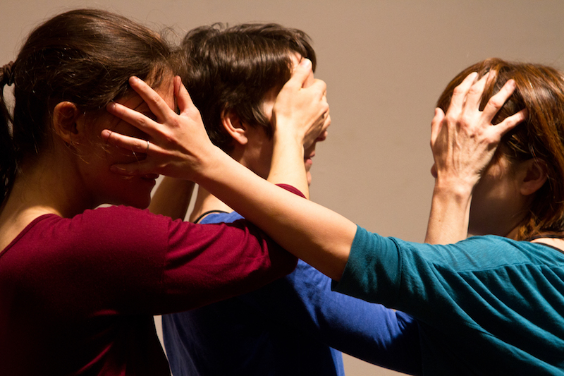 Three women in bright colored shirts place their hands over one another's faces